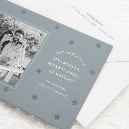 Close up of coastal blue custom holiday photo card paired with a black and white photo with white foil greeting 'may your days be heartfelt, meaningful, and bright' with dark blue snowflake pattern letterpress printed paired with a white envelope