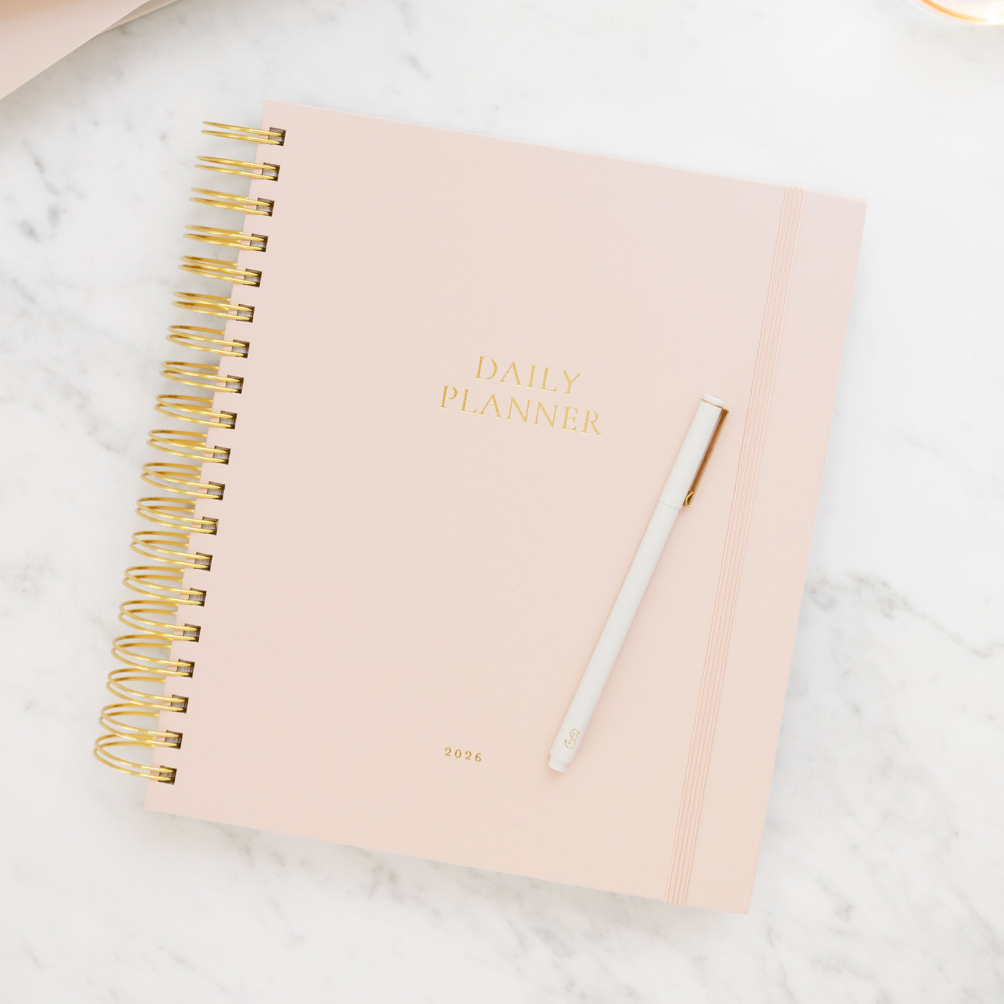Pink daily planner with gold spiral binding and pen on a marble surface