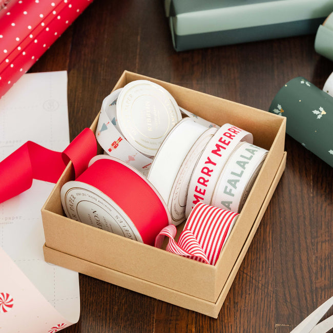 A kraft gift box filled with assorted decorative ribbons on a dark wooden table. The ribbons include solid red, white with red text, and red-and-white striped patterns.