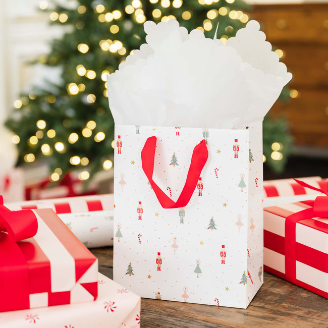 A white Christmas-themed gift bag filled with white tissue paper, decorated with red handles and a festive print of candy canes, trees, and nutcrackers, set on a wooden table with wrapped gift boxes and a blurred Christmas tree in the background.
