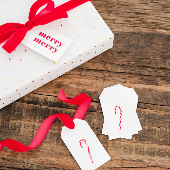 White gift wrapping paper with small gold polka dots, wrapped around a rectangular box and tied with a bright red ribbon. A white gift tag with red 'merry merry' text is attached, and two more matching white tags with red candy cane prints are placed on a rustic wooden surface.