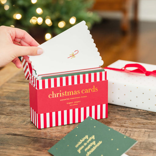 A hand is opening a red and white striped box of Christmas cards on a wooden table, with a blurred Christmas tree in the background and a wrapped gift box nearby.