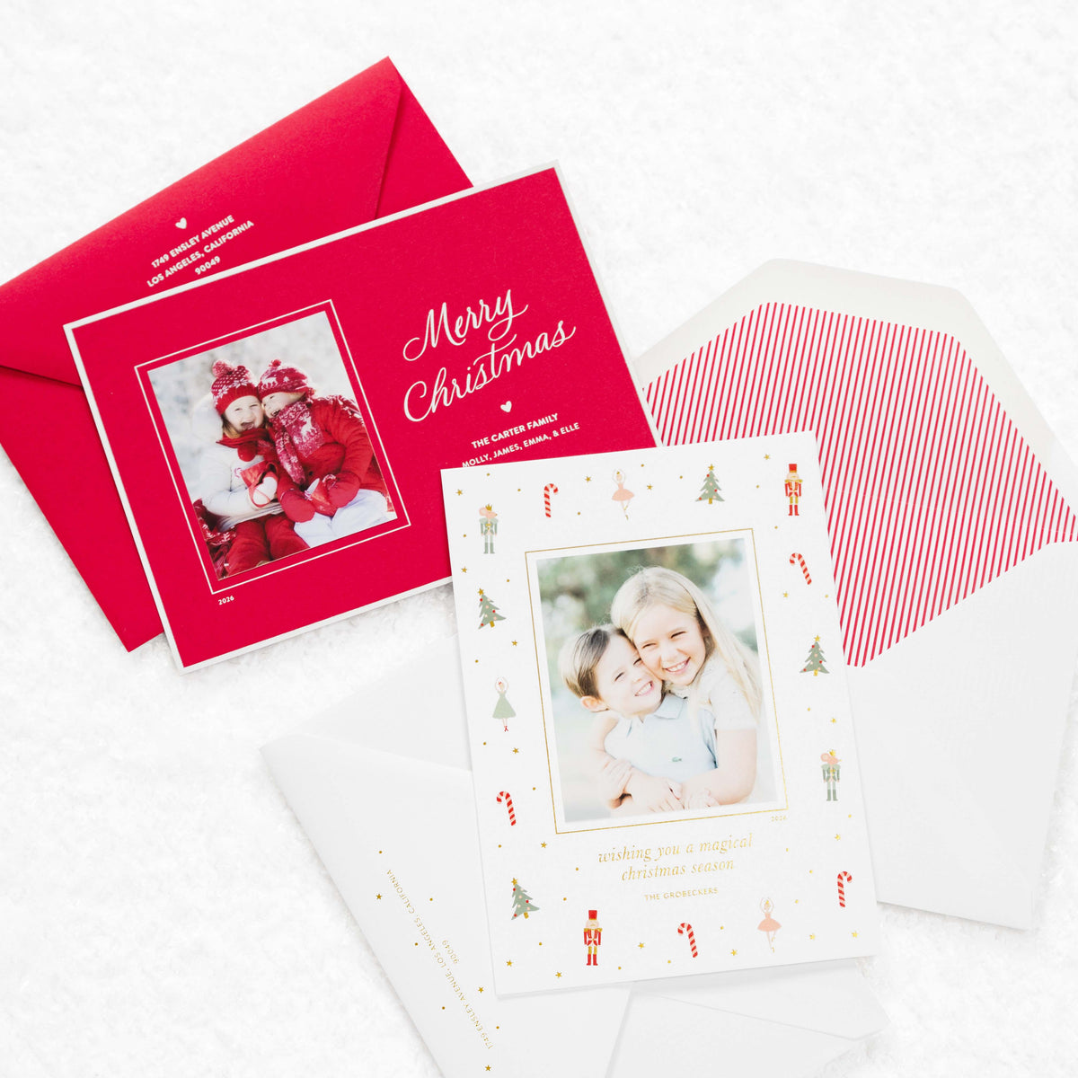 A styled flat-lay of personalized Christmas greeting cards and matching envelopes on a white textured background. One red envelope shows the front with a photo of children in red holiday clothes and the words 'Merry Christmas'; another white card with a photo of a smiling children is surrounded by small Christmas illustrations and gold foil accents, with a red-and-white striped inner envelope visible behind it.