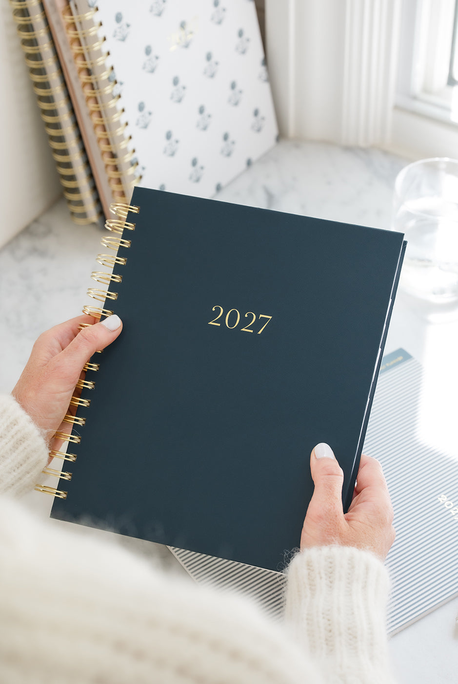 A woman holds a navy planner with "2027" printed on the cover in gold foil.