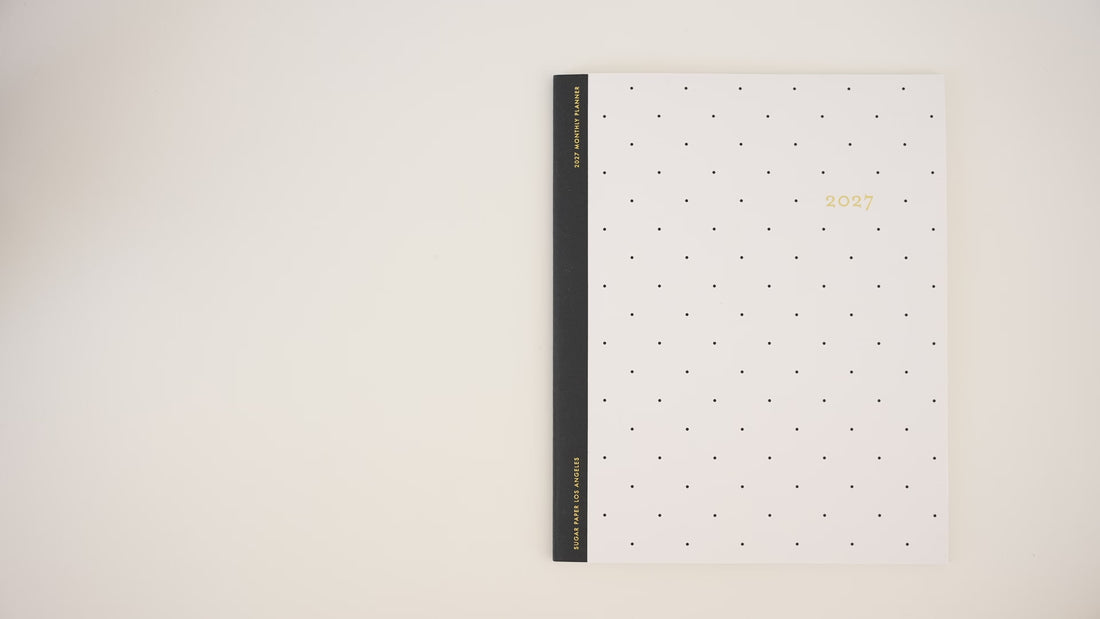A video of someone opening the flat monthly 2027 planner with a white with gold dot pattern and black tape binding printed with '2027'