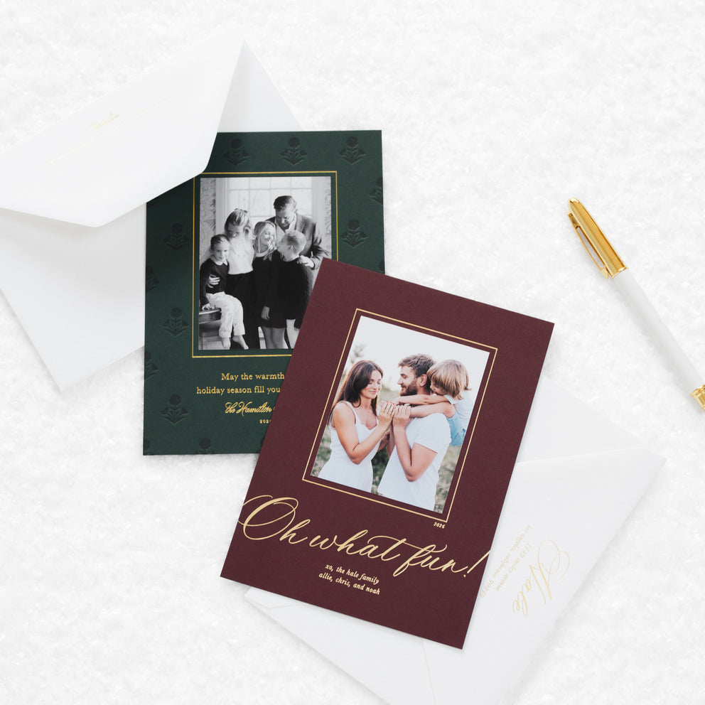 Two custom family holiday photo cards styled on a white textured background with white envelopes and a gold pen. The foreground card is burgundy with gold foil script reading 'Oh what fun!' and a family photo.