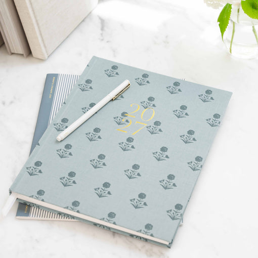 A light blue patterned planner with gold foil '2027' on the cover, styled on a white marble surface with a white pen resting on top.