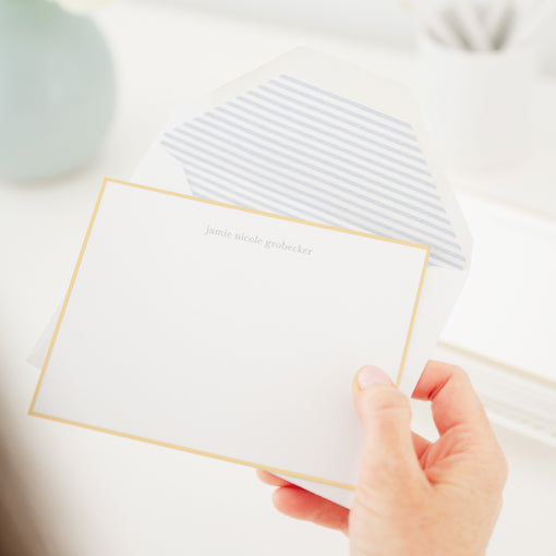 Hand holding a white card with gold border on a light background