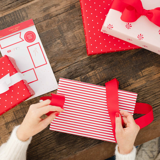 A top-down view of hands wrapping a gift box with red and white striped paper and a red ribbon, on a rustic wooden table. Other wrapped presents and a sheet of gift tags are visible in the background.
