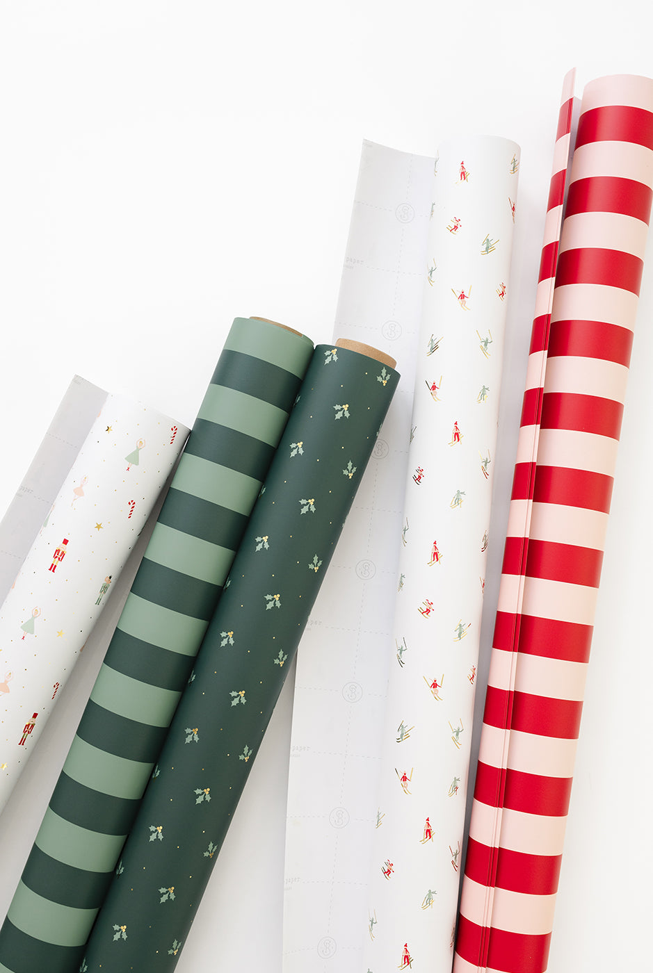 Several rolls of wrapping paper are arranged diagonally on a clean white background. The visible patterns include dark green and light green stripes, a dark green base with small white holly motifs, a white roll with tiny red and green holiday prints, and a bold red and white striped roll.