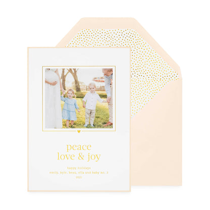 White custom holiday photo card printed with "peace love and joy" in gold foil with a photo of two kids and parents with a pink card border paired with a pink envelope open to gold teensy dot envelope liner