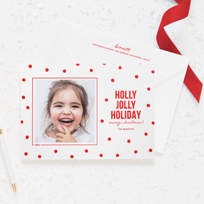 White holiday photo card printed with red dot pattern and 'holly jolly holiday merry christmas' in red ink paired with a white envelope with red return address
