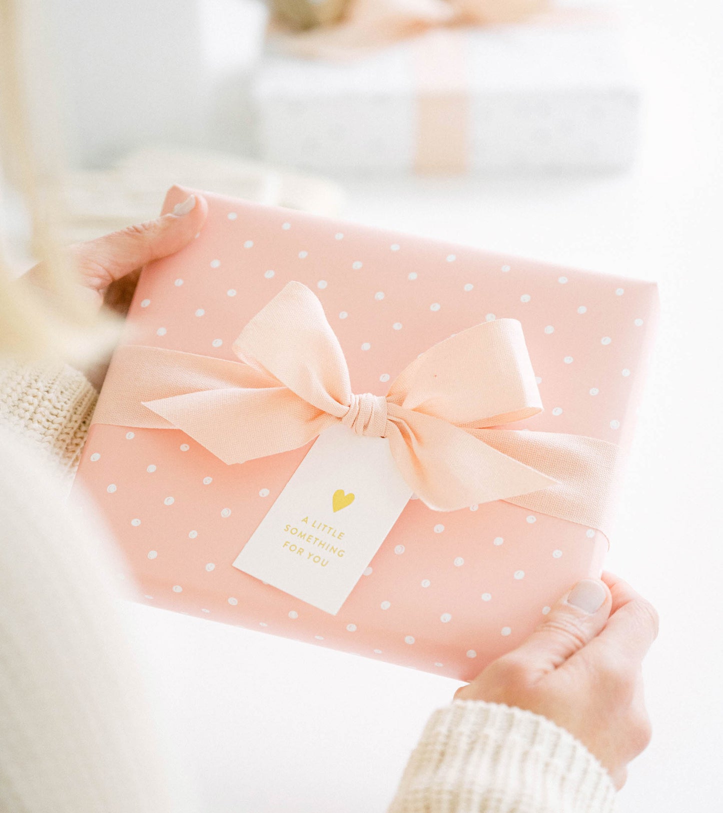 person holding gift wrapped in rose dot gift wrap with pale pink bow, and white gift tag printed with gold foil "a little something for you" with gold heart